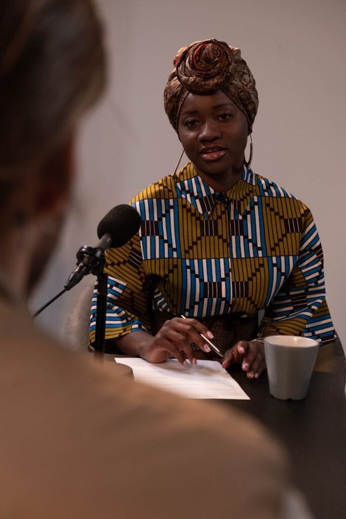 pexels-photo-6878721 Black woman in vibrant attire hosting a podcast interview, talking into a microphone.